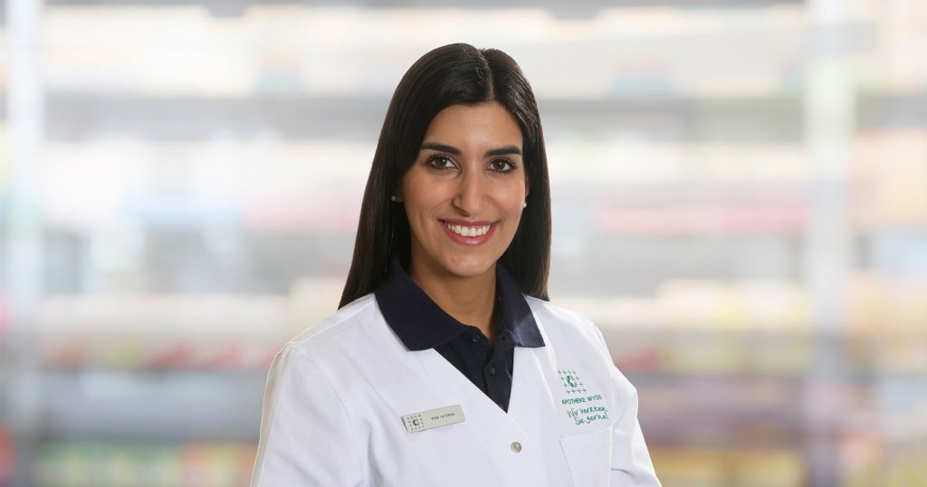 Caterina Vona, eine Frau mit langen dunklen Haaren, die einen weißen Laborkittel und ein marineblaues Hemd mit Kragen trägt, lächelt in die Kamera. Sie steht vor unscharfen Regalen, möglicherweise in einer Apotheke oder einem medizinischen Bereich.