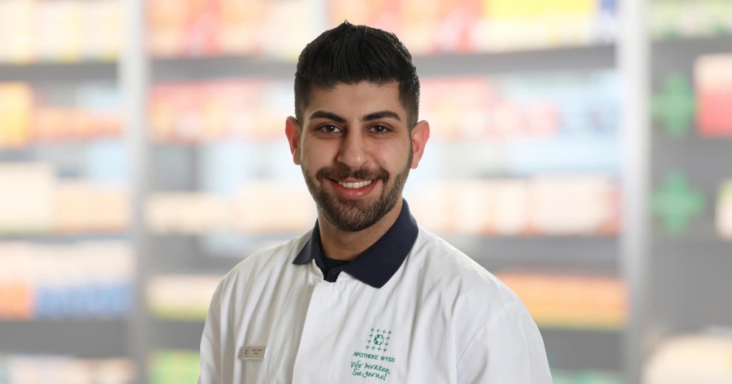 Gabriel Kouria, ein lächelnder Mann mit dunklem Haar und Bart, trägt einen weißen Laborkittel mit einem Namensschild und steht vor verschwommenen Apothekenregalen.