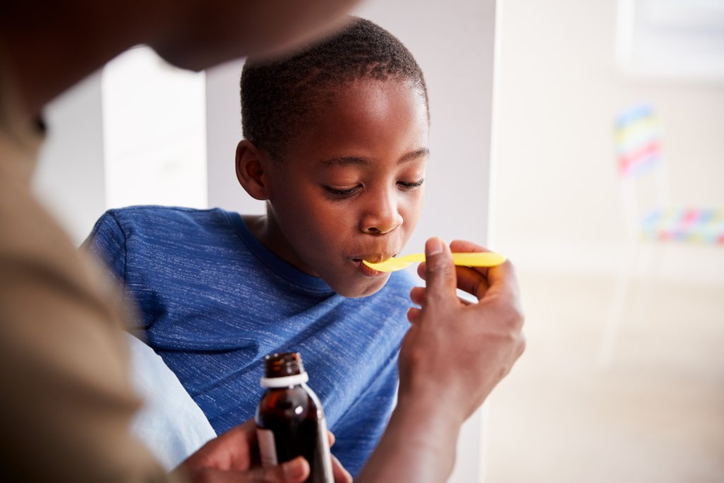 Ein Kind in einem blauen Hemd nimmt bei Kinderdosierungen Medizin aus einem gelben L&ouml;ffel, den ein Erwachsener h&auml;lt, der auch eine braune Medizinflasche in der Hand h&auml;lt. Das Kind scheint die Medizin zu schlucken.