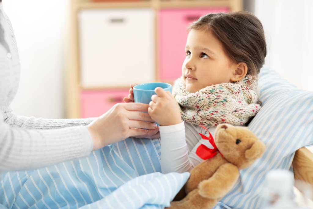 Ein kleines Kind mit einem Schal um den Hals sitzt im Bett und h&auml;lt einen blauen Becher in der Hand, w&auml;hrend ein Erwachsener ihn ihm reicht und f&uuml;r die richtige Kinderdosierung sorgt. Ein Teddyb&auml;r liegt neben ihr auf der Decke in dem gem&uuml;tlichen Schlafzimmer.