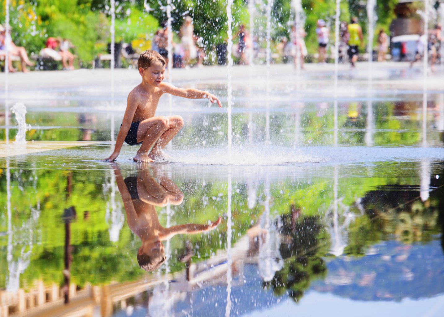 Ein kleiner Junge in Badehose hockt und spielt vergn&uuml;gt in einem Springbrunnen, der Hitze von &uuml;ber 30 Grad entfliehend, umgeben von Wasserstrahlen und Spiegelungen, mit verschwommenen Menschen und Gr&uuml;n im Hintergrund.