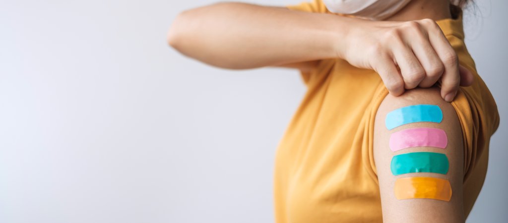 Eine Person, die ein gelbes Hemd und eine Gesichtsmaske tr&auml;gt, hebt ihren &Auml;rmel an und zeigt vier bunte Verb&auml;nde - blau, rosa, gr&uuml;n und orange - auf ihrem Oberarm, was auf mehrere COVID-19-Impfungen oder -Injektionen schlie&szlig;en l&auml;sst.