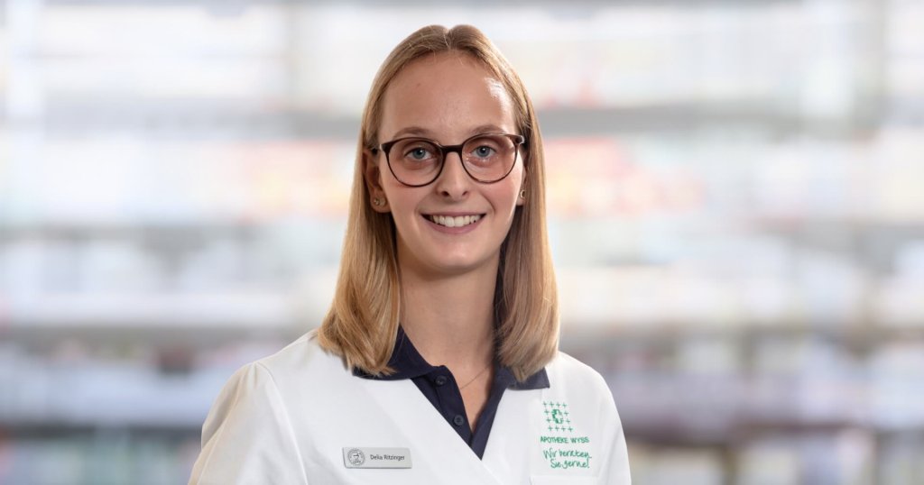 Delia Ritzinger, eine junge Frau mit glattem blondem Haar und Brille, lächelt in die Kamera. Sie trägt einen weißen Laborkittel und ein Namensschild und steht vor einem unscharfen Hintergrund.