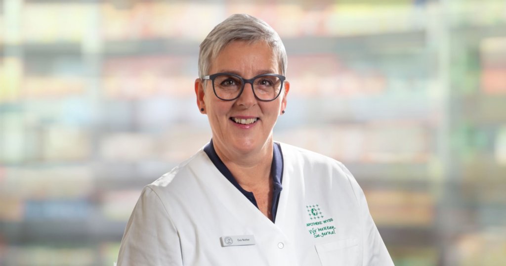 Eine lächelnde Frau mit kurzen grauen Haaren und Brille, die einen weißen Arztkittel trägt, steht vor einem unscharfen Hintergrund.