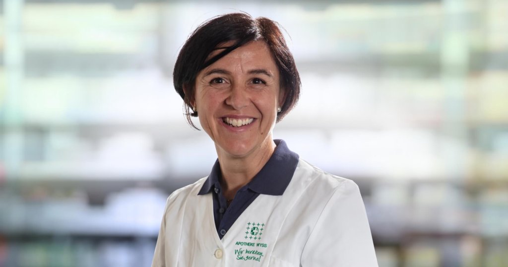 Karin Bearth Blumenthal, eine lächelnde Frau mit kurzen dunklen Haaren, trägt einen weißen Laborkittel über einem marineblauen Kragenhemd und steht vor einem unscharfen Hintergrund, der an Regale in einer Apotheke oder einem Labor erinnert.