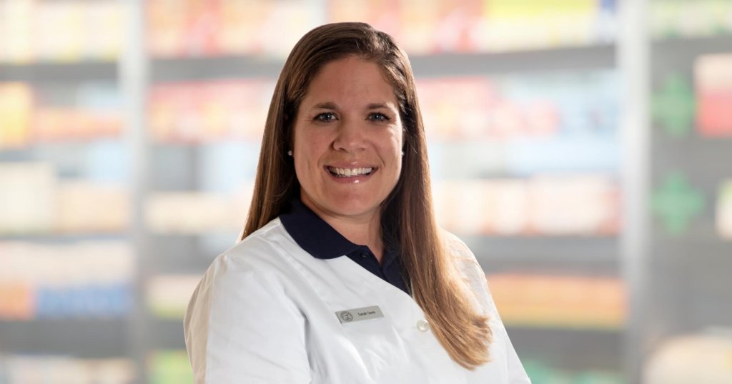 Eine Frau mit langen braunen Haaren, die einen weißen Laborkittel und ein Namensschild trägt, lächelt in die Kamera. Der Hintergrund scheint aus Regalen mit Produkten zu bestehen, wahrscheinlich in einer Apotheke oder einem Geschäft.