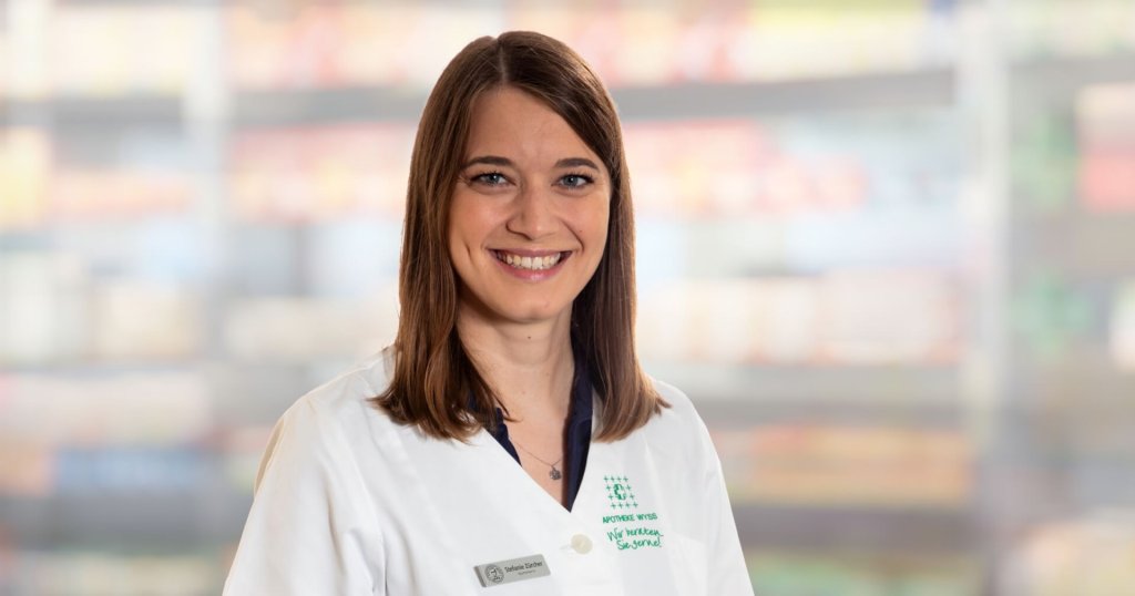 Stefanie Zürcher, eine Frau mit glattem braunem Haar und weißem Laborkittel, lächelt in die Kamera. Der Hintergrund ist unscharf, mit weichen Farben und undeutlichen Formen, die auf einen Innenraum hindeuten.