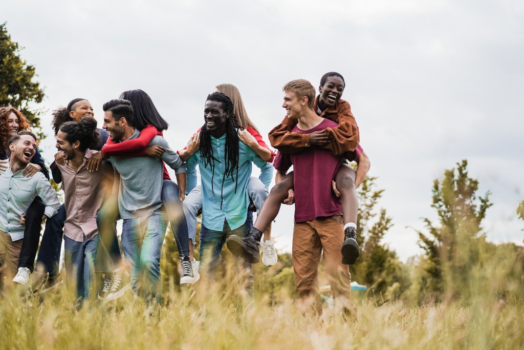 Eine Gruppe fr&ouml;hlicher junger Erwachsener spielt Huckepack in einem grasbewachsenen Park, lacht und l&auml;chelt zusammen unter einem bew&ouml;lkten Himmel. B&auml;ume und Gr&uuml;nfl&auml;chen f&uuml;llen den Hintergrund und schaffen eine lebendige Szene im Freien, die perfekt zu das Immunsystem st&auml;rken passt.