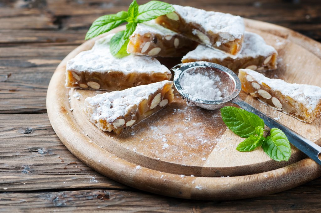 Aufgeschnittene Nuss- und Trockenfrüchtekuchen mit Puderzucker bestäubt auf einem Holzbrett, begleitet von einem Metallsieb und frischen Minzblättern, präsentieren die hausgemachten Leckereien auf einer rustikalen Holzoberfläche.