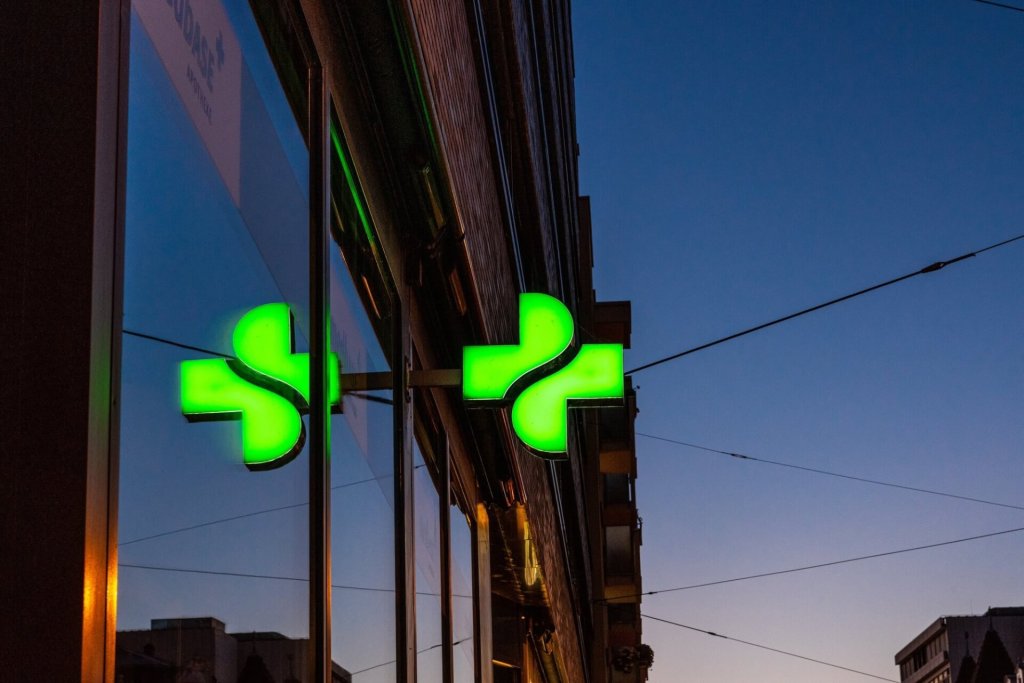 Ein leuchtend grünes Apothekenkreuz mit der Aufschrift "Tag und Nacht" ist in der Abenddämmerung an einem Gebäude angebracht und spiegelt sich im Fenster. Stromleitungen und Silhouetten von Gebäuden heben sich vom Abendhimmel im Hintergrund ab.
