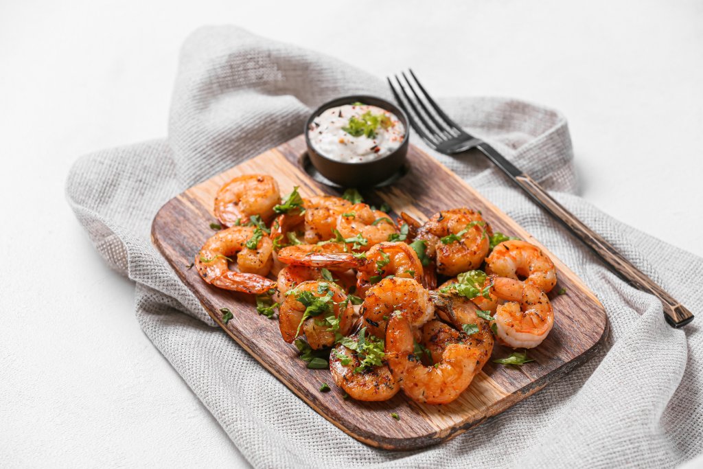 Gegrillte Garnelen, garniert mit gehackten Kr&auml;utern, serviert auf einem Holzbrett, begleitet von einem Sch&auml;lchen mit Dip-Sauce. Besonders ideal f&uuml;r Heuschnupfengeplagte, da Garnelen selten mit Kreuzallergie in Verbindung gebracht werden. Eine Gabel liegt auf einer grauen Serviette neben dem Brett.