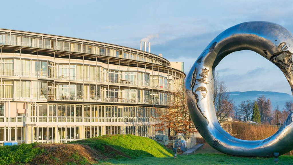 Ein gro&szlig;es, modernes Geb&auml;ude mit vielen Fenstern steht neben einer metallisch gl&auml;nzenden, ringf&ouml;rmigen Skulptur auf einer Wiese, wo G&auml;rtnern als eine Form der Therapie verstanden wird, umgeben von B&auml;umen und Bergen unter einem klaren Himmel.