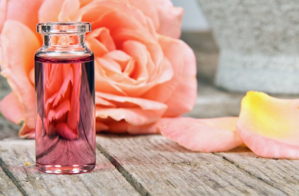 Ein kleines, mit rosa Fl&uuml;ssigkeit gef&uuml;lltes Glasfl&auml;schchen steht auf einer h&ouml;lzernen Oberfl&auml;che und erinnert an die Heilkraft der D&uuml;fte. Im Hintergrund weisen eine zartrosa Rose und verstreute Bl&uuml;tenbl&auml;tter auf die wohltuende Wirkung der Aromatherapie hin.