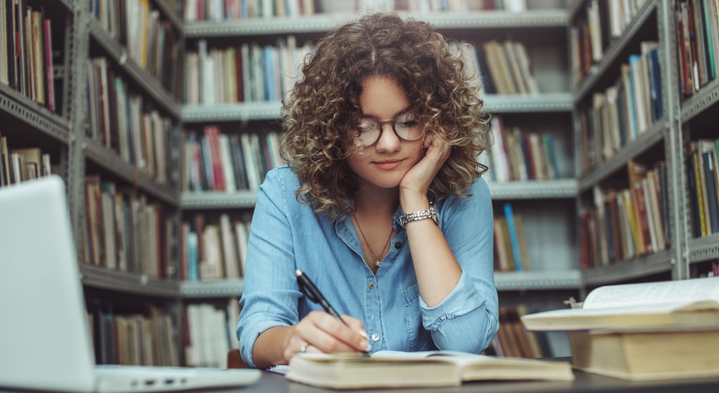 Eine junge Frau mit lockigem Haar und Brille sitzt an einem Tisch in einer Bibliothek und liest &uuml;ber M&ouml;glichkeiten, die Gehirnleistung zu steigern, und macht sich Notizen in einem Notizbuch, umgeben von aufgeschlagenen B&uuml;chern &uuml;ber Nootropika und ihrem Laptop. Im Hintergrund stehen B&uuml;cherregale.