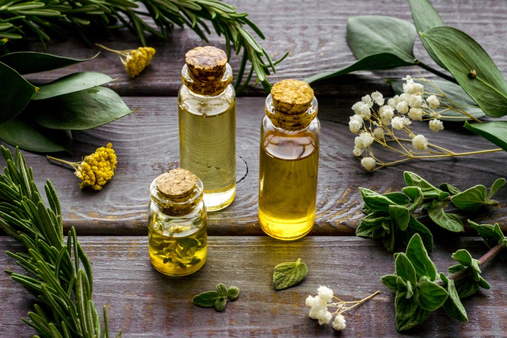 Drei kleine Glasflaschen mit Korkstopfen, gefüllt mit gelben und klaren Ölen, umgeben von frischen grünen Kräutern und kleinen weißen und gelben Blumen auf einer rustikalen Holzoberfläche - perfekt für Aromatherapie und die Heilkraft wohltuender Düfte.