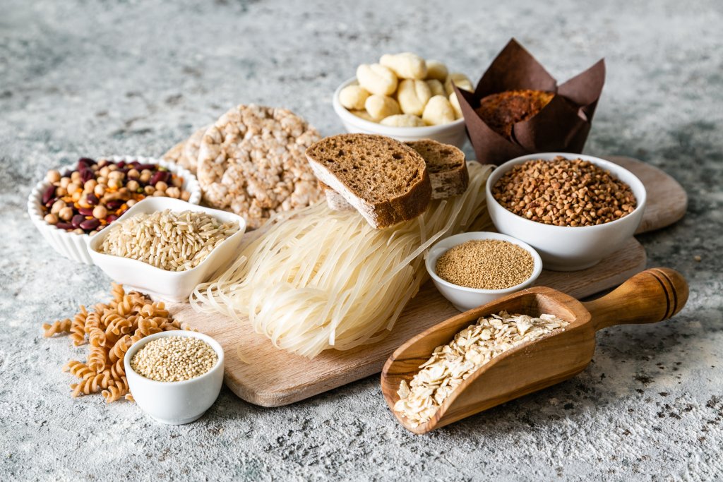 Eine Vielzahl von glutenfreien Lebensmitteln für Menschen mit Glutenunverträglichkeit oder Zöliakie, darunter Reisnudeln, Schwarzbrot, Bohnen, Hafer, Buchweizen, Hirse, Puffreiskuchen, Gnocchi und Muffins - hübsch angerichtet auf Holzbrettern und in Schalen.
