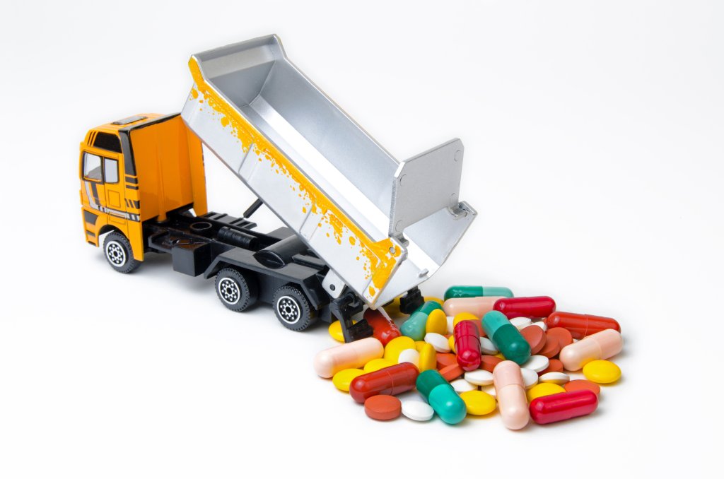 Ein gelber Spielzeugkipper wird gezeigt, wie er einen Haufen bunter Pillen, Tabletten und Kapseln auf eine wei&szlig;e Fl&auml;che abl&auml;dt.