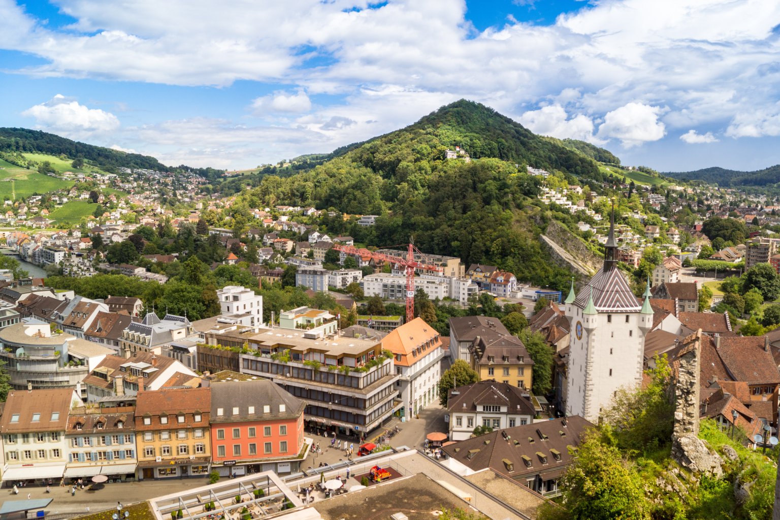 Das Panorama einer Schweizer Stadt mit farbenfrohen Gebäuden, einem Kirchturm mit Uhr und üppig grünen Hügeln und Bergen im Hintergrund unter einem teilweise bewölkten Himmel fängt den Geist des 100-jährigen Jubiläums der Badenfahrt ein.