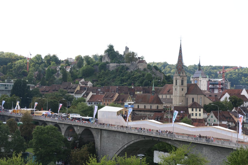 Eine steinerne Br&uuml;cke, &uuml;ber die Menschen spazieren, &uuml;berspannt ein gr&uuml;nes Tal; dahinter erheben sich in der N&auml;he eines Schlosses auf einem H&uuml;gel historische Geb&auml;ude und eine Kirche mit einem hohen Turm, alles Teil der Feierlichkeiten zum 100-j&auml;hrigen Jubil&auml;um von Badenfahrt inmitten von B&auml;umen und Baukr&auml;nen.
