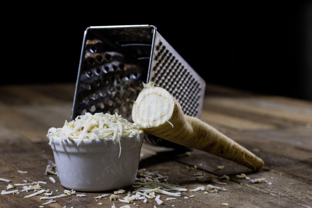 Eine Metallreibe liegt auf der Seite neben einer wei&szlig;en Sch&uuml;ssel mit frisch geriebenem Meerrettich, daneben liegt eine ganze Meerrettichwurzel auf der Holzfl&auml;che.