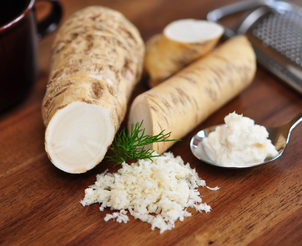 Ein Holztisch mit ganzen Meerrettichwurzeln, ein Haufen frisch geriebener Meerrettich, ein Löffel cremiger Soße, ein Dillzweig und eine Metallreibe im Hintergrund.