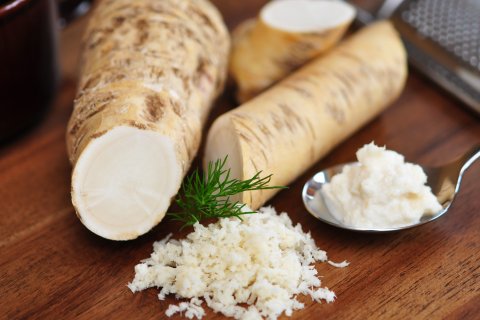 Ein Holztisch mit ganzen Meerrettichwurzeln, ein Haufen frisch geriebener Meerrettich, ein L&ouml;ffel cremiger So&szlig;e, ein Dillzweig und eine Metallreibe im Hintergrund.