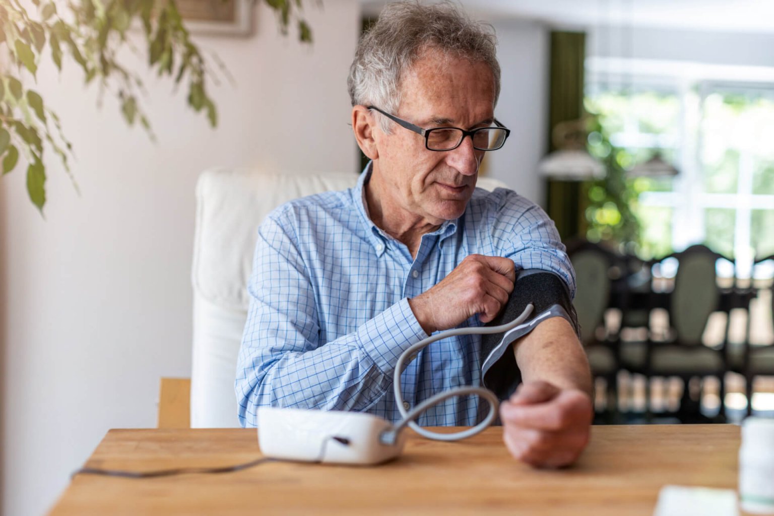 Ein &auml;lterer Mann mit Brille und blau-kariertem Hemd sitzt an einem Tisch und bedient ein elektronisches Blutdruckmessger&auml;t an seinem linken Arm in einem hellen, gem&uuml;tlichen Raum.