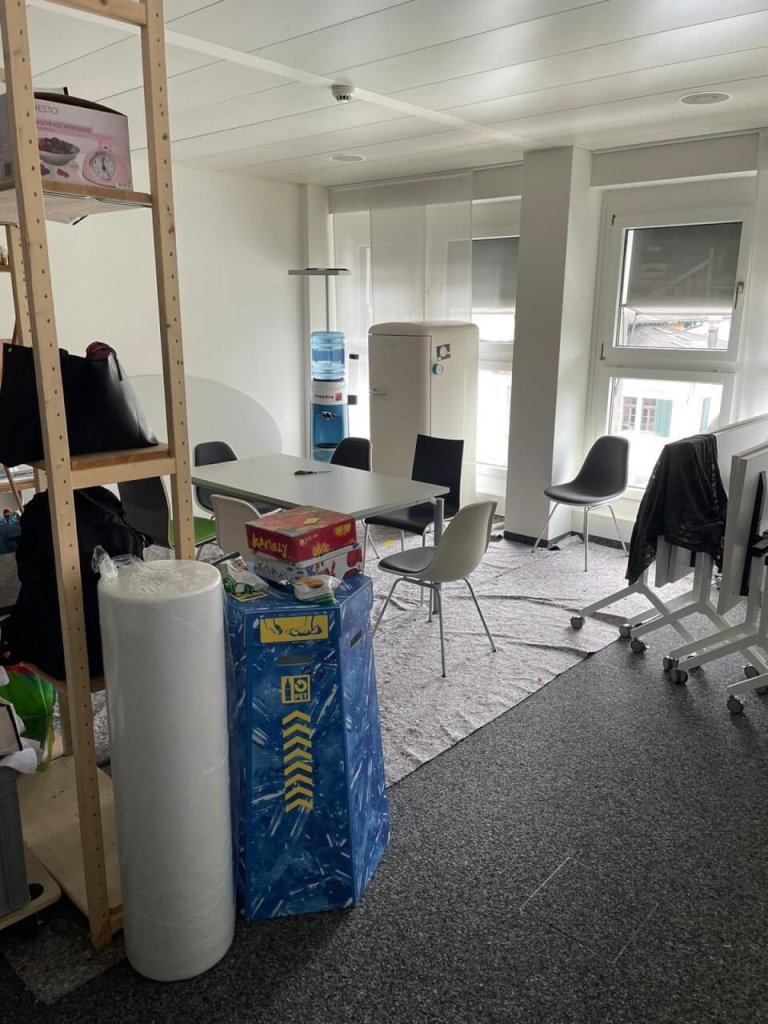 Ein kleiner Pausenbereich mit einem Tisch, vier St&uuml;hlen, einem Wasserspender und einem K&uuml;hlschrank. An einem Holzregal stapeln sich Brettspiele und Verbrauchsmaterialien, die die Kundenorientierung im Team unterst&uuml;tzen. Im Vordergrund steht Verpackungsmaterial; nat&uuml;rliches Licht str&ouml;mt herein.