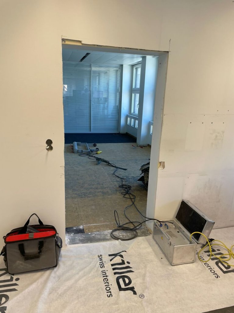 Eine teilweise konstruierte T&uuml;r&ouml;ffnung in einem B&uuml;ro mit offenen Kanten spiegelt einen kontinuierlichen Verbesserungsprozess wider. Bauwerkzeuge, Kabel und ein Werkzeugkasten liegen auf dem Boden. Eine wei&szlig;e Matte mit der Aufschrift "Kiler swiss interiors" bedeckt einen Teil des Bodens.