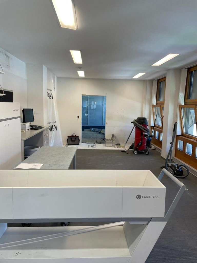 Ein Raum, der teilweise renoviert wird, mit verschiedenen Ger&auml;ten, einem CareFusion-Ger&auml;t im Vordergrund, einem Schreibtisch mit Monitoren auf der linken Seite, Baumaterialien an der R&uuml;ckwand und Fenstern auf der rechten Seite, die den kontinuierlichen Verbesserungsprozess widerspiegeln.