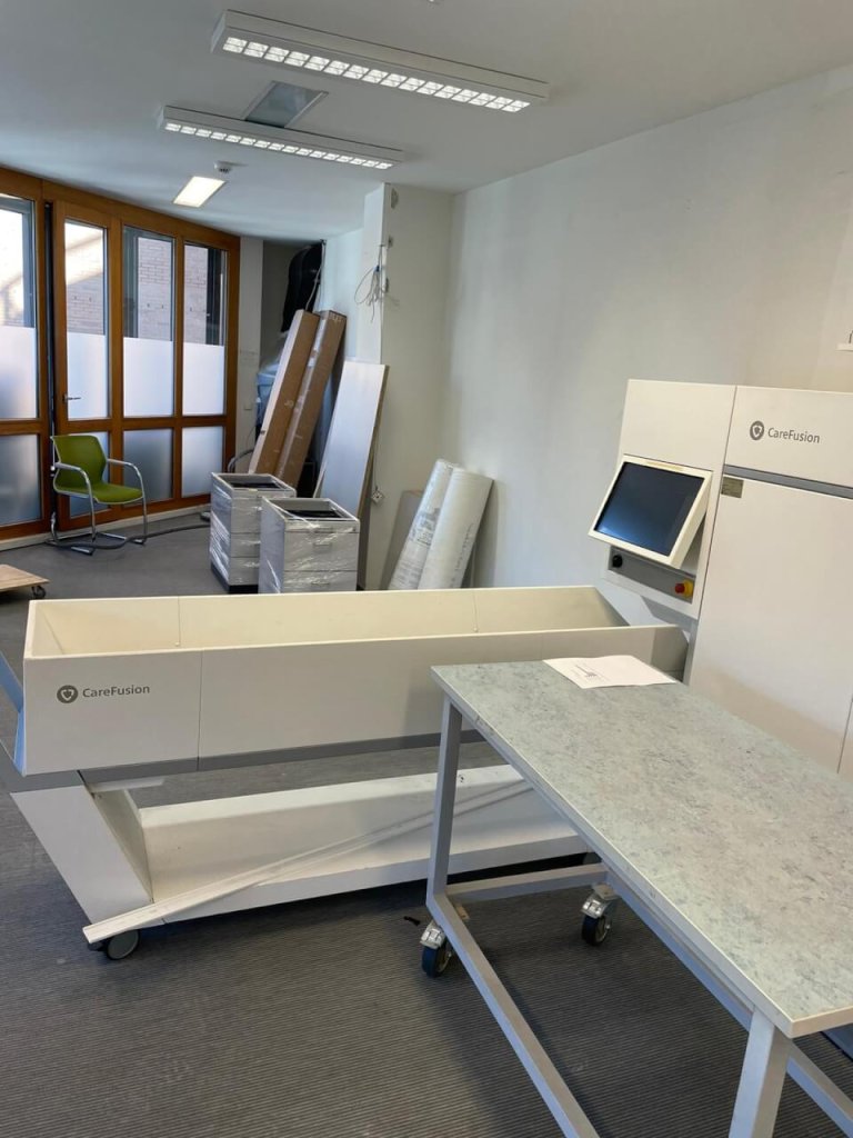 Ein medizinisches Ger&auml;t mit der Aufschrift "CareFusion", das QMS und Kundenorientierung unterst&uuml;tzt, steht neben einem Monitor und einem Rolltisch in einem hellen Raum mit gro&szlig;en Fenstern, gestapelten Kisten, einem gr&uuml;nen Stuhl und verpackten Gegenst&auml;nden an der Wand.