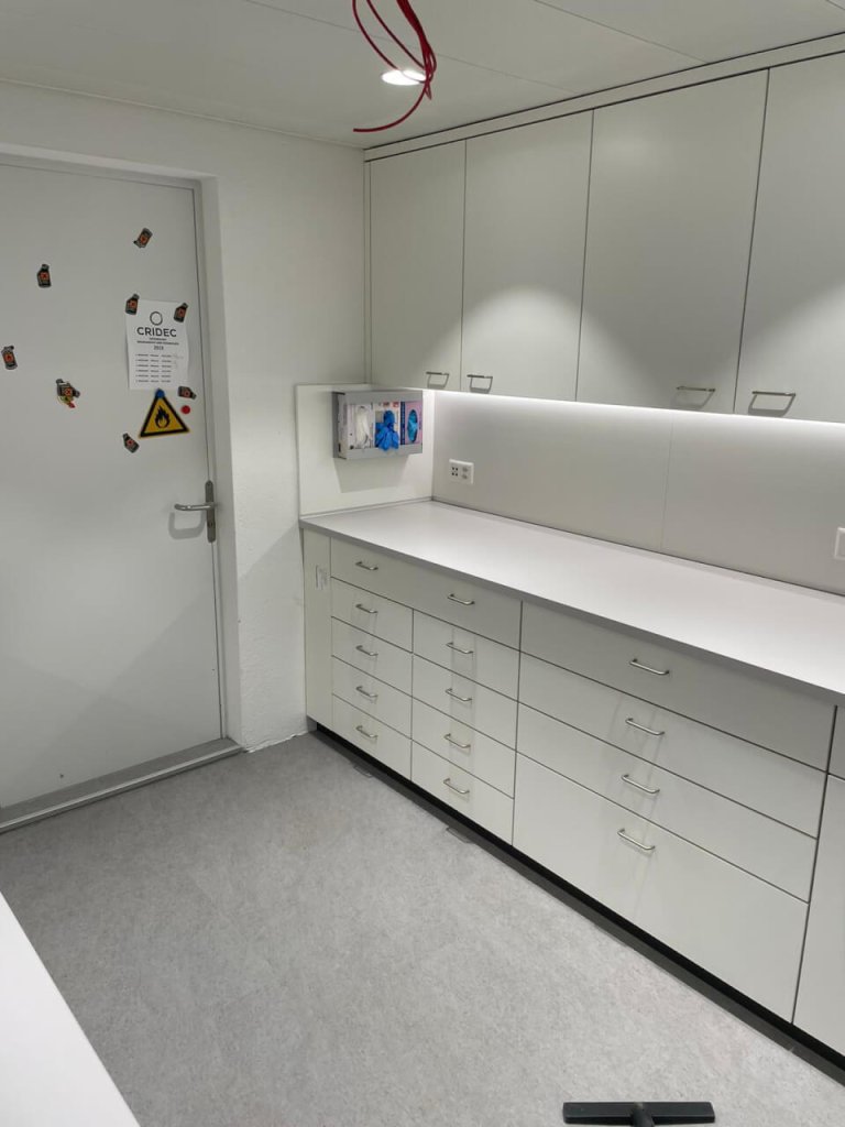 Ein sauberer, moderner medizinischer Raum mit wei&szlig;en Schr&auml;nken, Schubladen und einem an der Wand montierten Handschuhspender spiegelt eine sterile, organisierte Atmosph&auml;re wider, die von QMS-Grunds&auml;tzen und einem kontinuierlichen Verbesserungsprozess gepr&auml;gt ist.