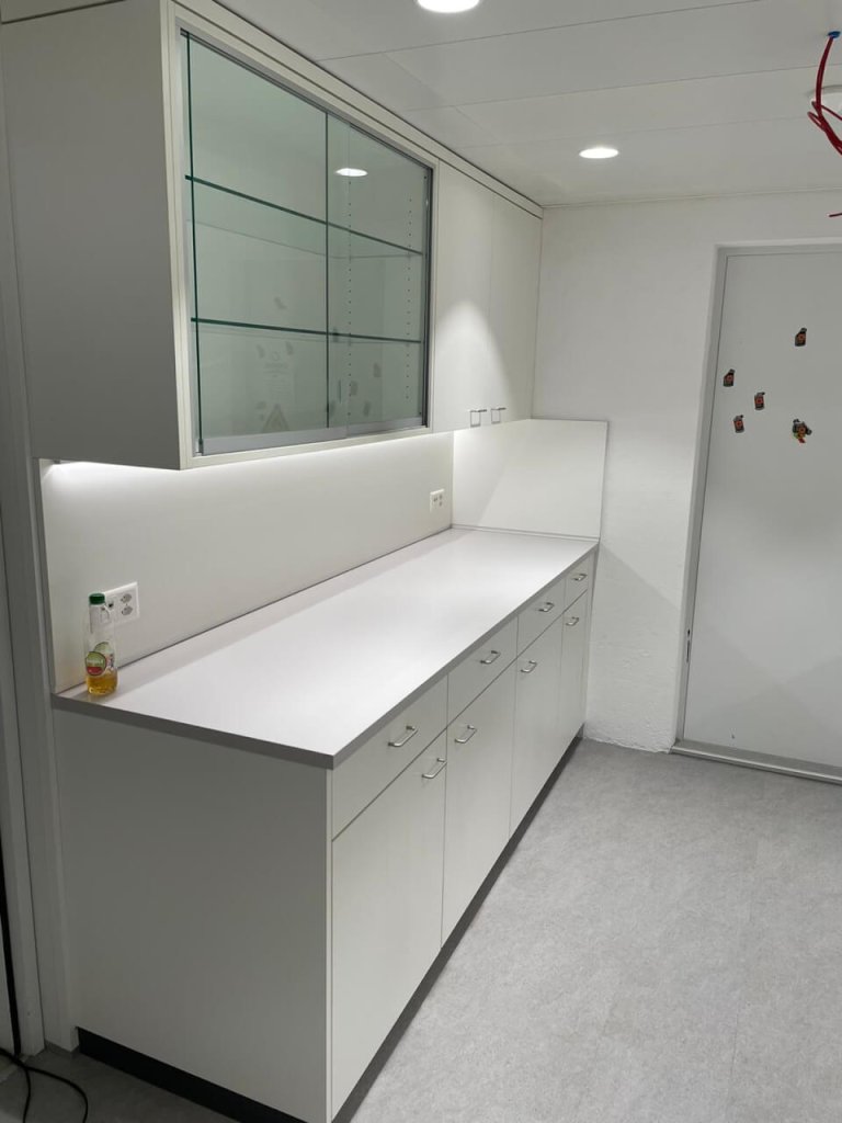 Eine saubere, moderne K&uuml;che mit wei&szlig;en Schr&auml;nken, Schubladen und Arbeitsfl&auml;chen spiegelt Kundenorientierung wider. Eine kleine Flasche steht auf dem Tresen unter einer Glasvitrine. Der Raum hat wei&szlig;e W&auml;nde und eine T&uuml;r mit Magneten.