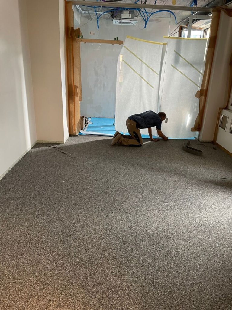Eine Person kniet auf einem neu verlegten Teppichboden in der N&auml;he einer T&uuml;r, die mit abgeklebten Plastikplanen in einem im Bau befindlichen Raum abgedeckt ist, und reflektiert die Kundenorientierung, w&auml;hrend sie sorgf&auml;ltig arbeitet. Rundherum sind Werkzeuge und QMS-bezogene Materialien zu sehen.