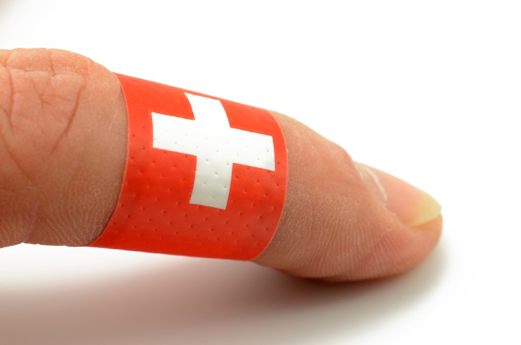 Nahaufnahme eines Fingers mit einem roten Pflaster mit einem wei&szlig;en Kreuz, das Gesundheit und Erste Hilfe symbolisiert, um die Fingerkuppe gewickelt. Der schlichte wei&szlig;e Hintergrund unterstreicht den Fokus auf Wohlbefinden und Pflege.