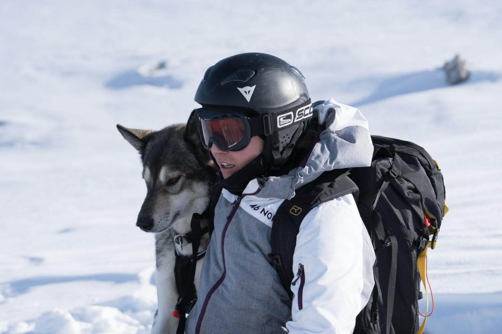 Eine Person in Winterausr&uuml;stung und mit Helm steht im Schnee und lehnt sich an einen Husky Hund. Die helle, verschneite Szene zeigt beide, wie sie w&auml;hrend eines Einsatzes in der freien Natur eine Pause einlegen, und unterstreicht ihre Verbundenheit und Liebe zu Abenteuer und Gesundheit.