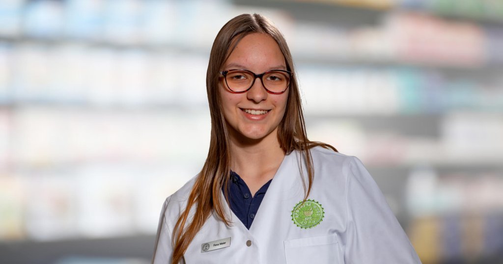 Eine junge Frau mit langen braunen Haaren und Brille lächelt, während sie einen weißen Apothekenkittel und ein Namensschild trägt und vor verschwommenen, mit Produkten gefüllten Regalen steht.