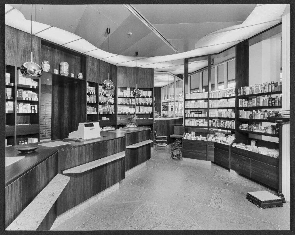 Ein Schwarz-Weiss-Foto des Interieurs der Apotheke Wyss am Bahnhof Baden zeigt schlichte Holzeinbauten, mit Medikamenten best&uuml;ckte Regale und eine alte Registrierkasse - eine perfekte Mischung aus Modernisierung und klassischem Apothekencharme in einem hellen, &uuml;bersichtlichen Raum.
