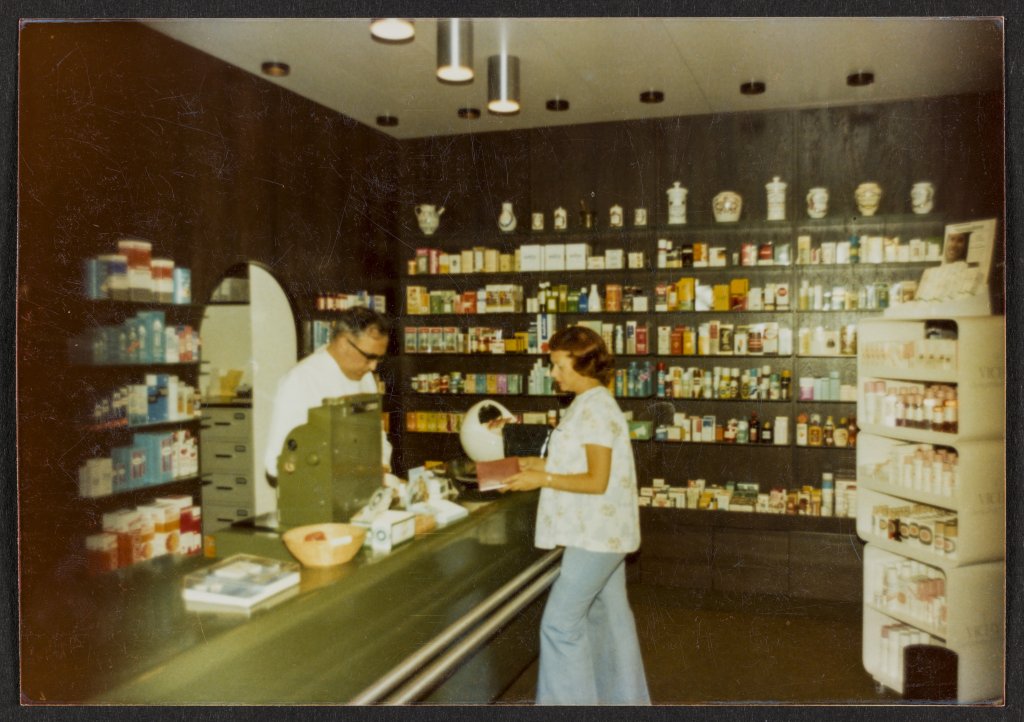 Eine Frau steht an der Theke der Apotheke Wyss und spricht mit einem Mann hinter der Kasse. Dahinter stehen Regale mit Medikamentenschachteln und Gesundheitsprodukten, die auf die Bahnhofsapotheke vor der Modernisierung in den 1970er Jahren hindeuten.