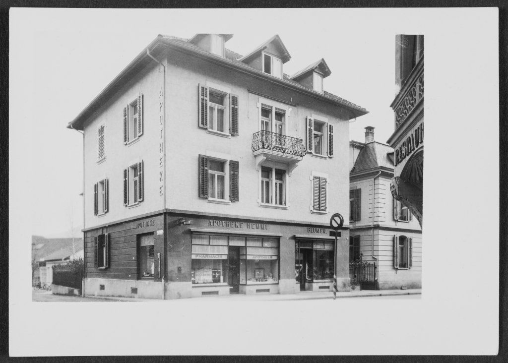 Schwarz-Weiss-Foto eines dreist&ouml;ckigen Eckgeb&auml;udes mit Fensterl&auml;den und einem kleinen Balkon. Im Erdgeschoss befindet sich die Apotheke Wyss mit grossen Schaufenstern und Beschriftungen in der N&auml;he des Bahnhofs Baden.