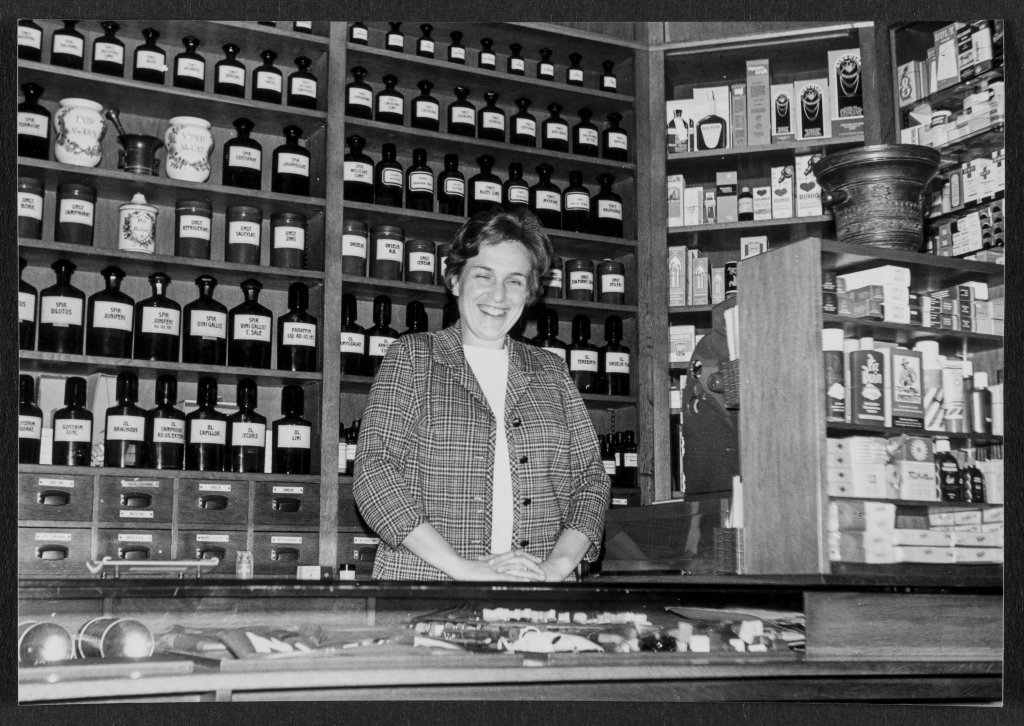 Eine Frau l&auml;chelt hinter dem Tresen der alten Apotheke Wyss, umgeben von etikettierten Flaschen und Gl&auml;sern. Die altmodische Apotheke in der N&auml;he des Bahnhofs Baden bewahrt die Tradition und setzt gleichzeitig auf behutsame Modernisierung.