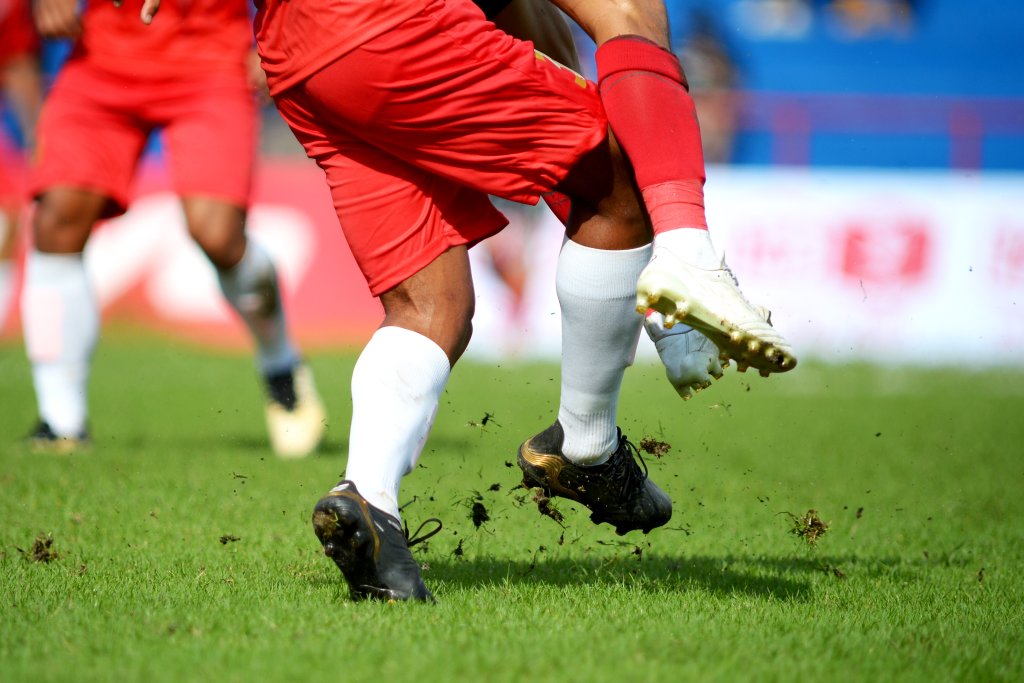Nahaufnahme von zwei Fußballspielern in roten Shorts und weißen Stutzen, die auf einer Wiese um den Ball kämpfen, wobei Dreck aufgewirbelt wird, als der eine den anderen angreift - was sowohl den Nervenkitzel als auch das Risiko von Sportverletzungen ohne entsprechende Präventionstipps verdeutlicht.
