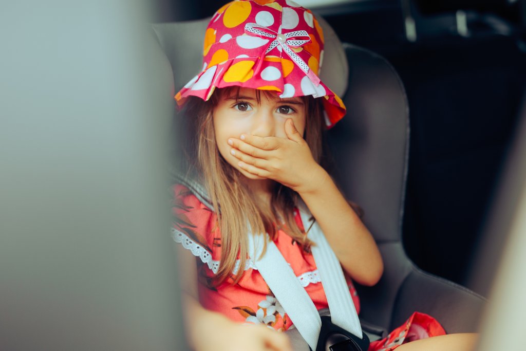 Ein junges Mädchen mit farbenfrohem Hut und rotem Kleid sitzt auf einem Autositz, hält sich den Mund mit der Hand zu und schaut mit großen Augen nach vorne - vielleicht spürt es die ersten Anzeichen einer Reisekrankheit.