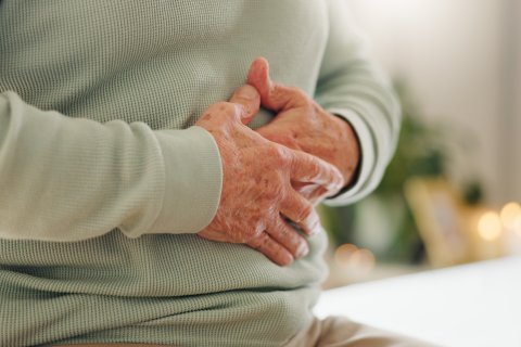 Eine Person, die einen hellgr&uuml;nen Pullover tr&auml;gt, sitzt und h&auml;lt sich mit beiden H&auml;nden den Bauch und scheint sich unwohl zu f&uuml;hlen oder Schmerzen zu haben - ein Bild, das h&auml;ufig in Interviews &uuml;ber Darmgesundheit diskutiert wird. Der Hintergrund ist sanft verschwommen.