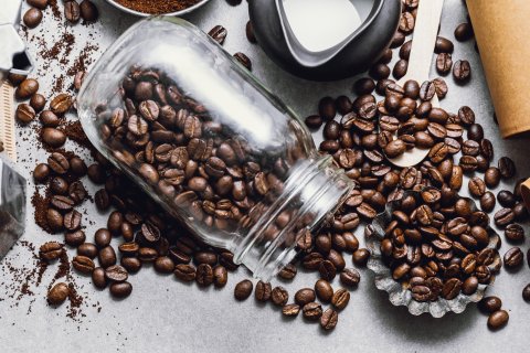 Ein Glasgef&auml;&szlig; versch&uuml;ttet Kaffeebohnen auf einen Tisch, der von einer Mokkakanne, einem L&ouml;ffel mit gemahlenem Kaffee, einer Schale mit Zucker, einem Milchk&auml;nnchen, braunem Zucker und zwei braunen Tassen zum Mitnehmen umgeben ist.