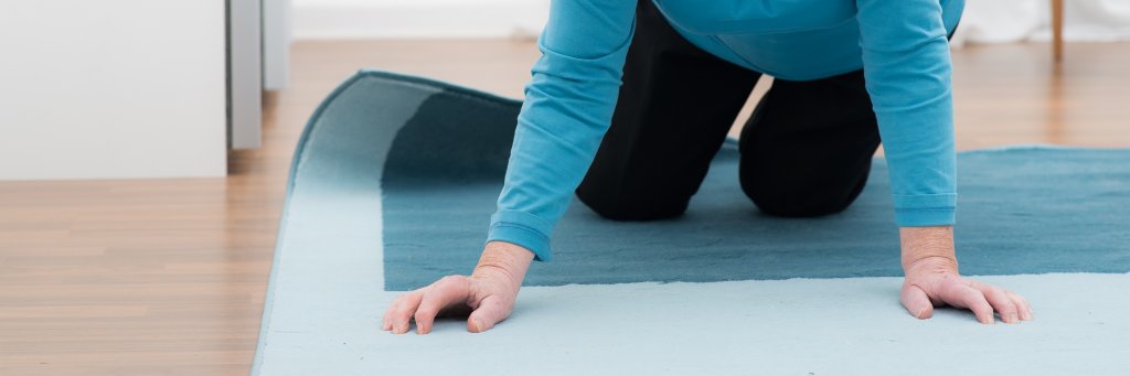 Ein älterer Erwachsener in blauem Hemd und dunkler Hose liegt auf Händen und Knien auf einem blauen Teppich und scheint sich vom Boden zu erheben, um die Bedeutung der Sturzprävention am Unfallort Zuhause zu verdeutlichen. Nur der Oberkörper und die Arme sind sichtbar.