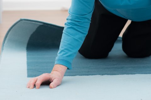 Ein &auml;lterer Erwachsener in blauem Hemd und dunkler Hose liegt auf H&auml;nden und Knien auf einem blauen Teppich und scheint sich vom Boden zu erheben, um die Bedeutung der Sturzpr&auml;vention am Unfallort Zuhause zu verdeutlichen. Nur der Oberk&ouml;rper und die Arme sind sichtbar.