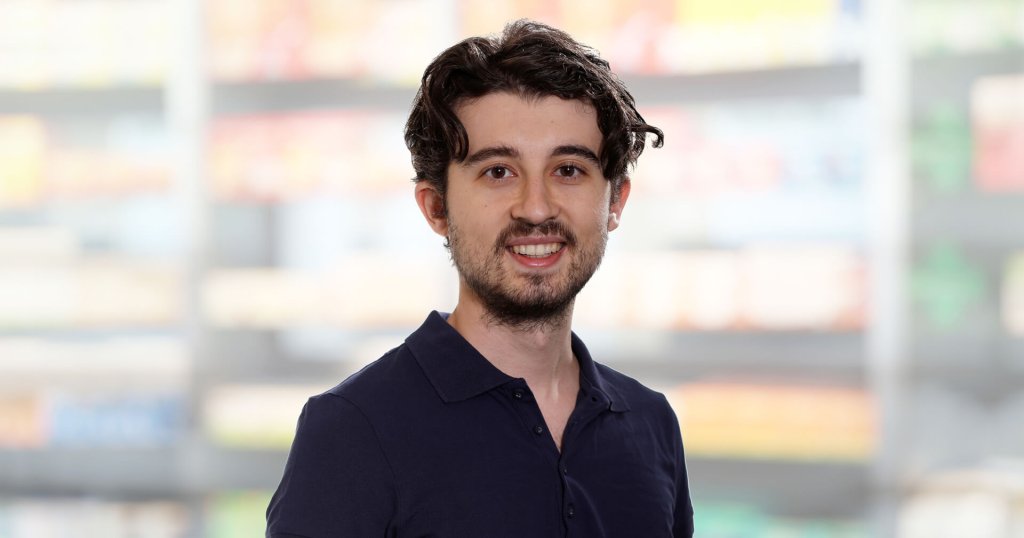 Ein junger Mann mit dunklem, gewelltem Haar und einem kurzen Bart lächelt in die Kamera und trägt ein marineblaues Polohemd. Der Hintergrund ist unscharf und zeigt Regale mit bunten Produkten, die an einen Laden oder Supermarkt erinnern.