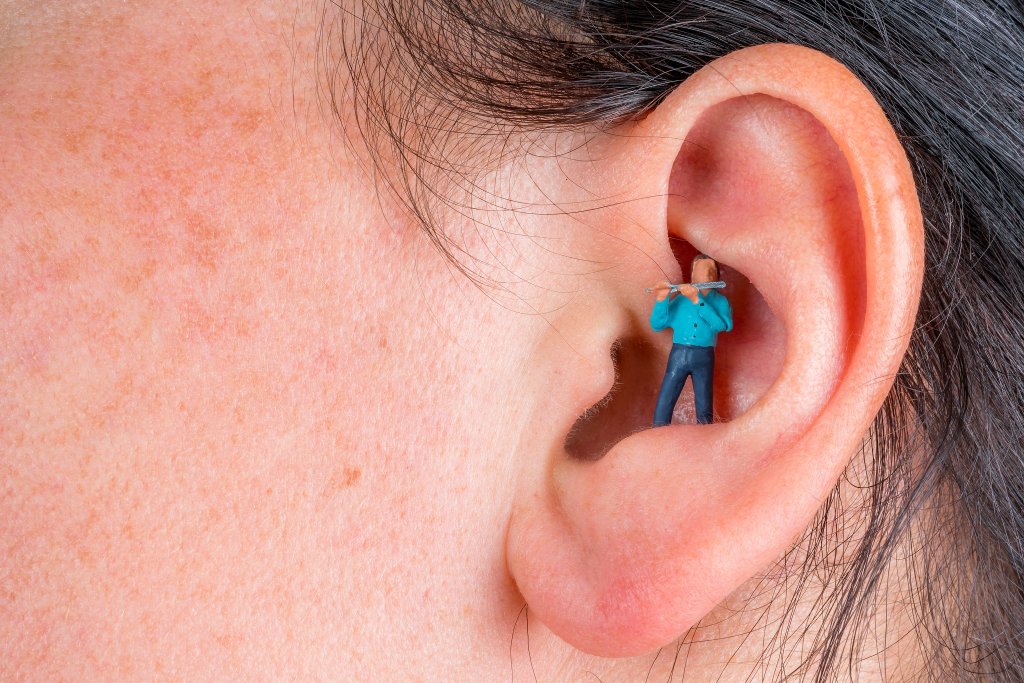 Eine Nahaufnahme eines menschlichen Ohrs mit einer digital hinzugefügten winzigen Figur in einem blauen Hemd, die im Inneren des Gehörgangs steht und ihn mit einem großen Wattestäbchen zu reinigen scheint - als Symbol für den Wunsch, Nebengeräusche aus dem täglichen Leben zu entfernen.