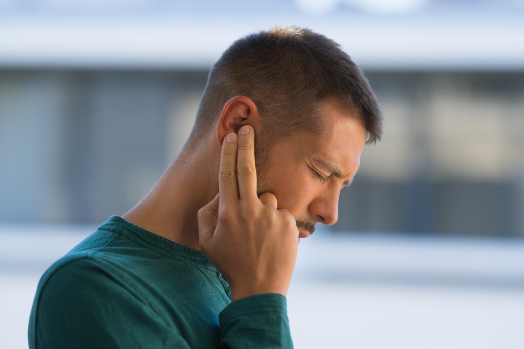 Ein Mann in einem gr&uuml;nen Hemd schlie&szlig;t die Augen und dr&uuml;ckt seine Finger auf sein Ohr. Er scheint sich unwohl zu f&uuml;hlen oder Schmerzen zu haben, m&ouml;glicherweise aufgrund eines Tinnitus. Der Hintergrund ist unscharf.